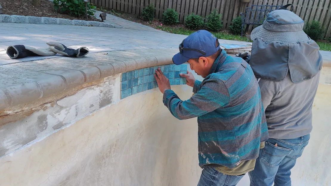 Workers installing teal mosaic tiles along a pool waterline